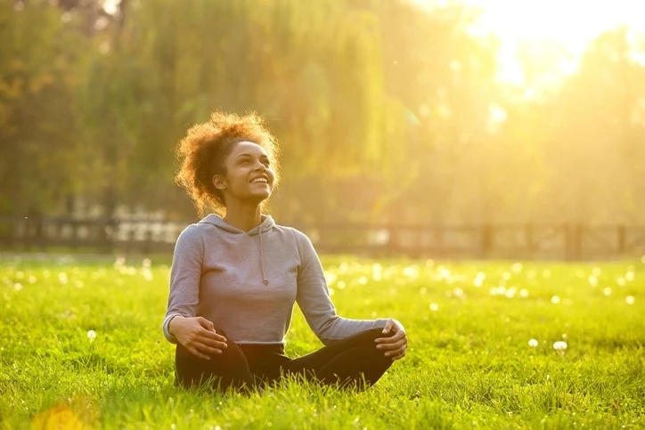 Smiling woman practicing meditation outdoors in a sunlit park, promoting stress relief and mindfulness techniques